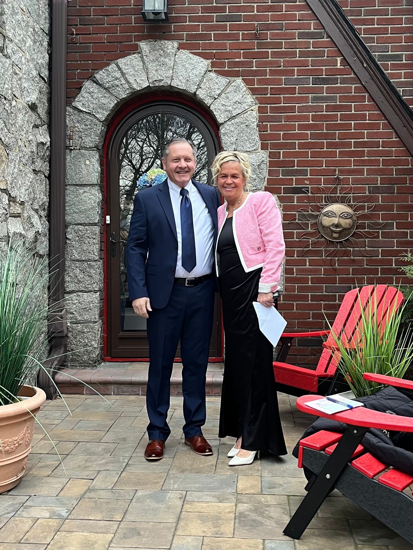Smiling couple in formal attire standing on a patio with red chairs and brick wall backdrop.